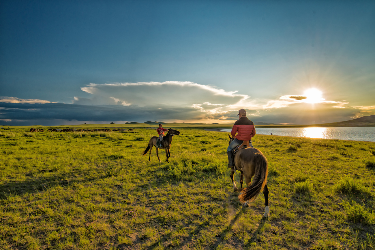 Two nomadic Mongolians riding their horses at sunset